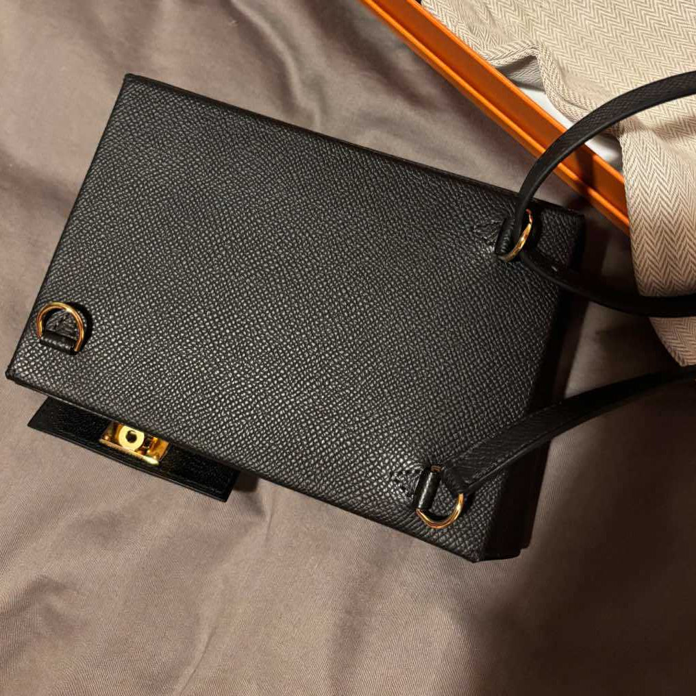 A black textured leather handbag with gold-tone hardware, shown next to its orange and white branded box and dust bag on a gray fabric surface.