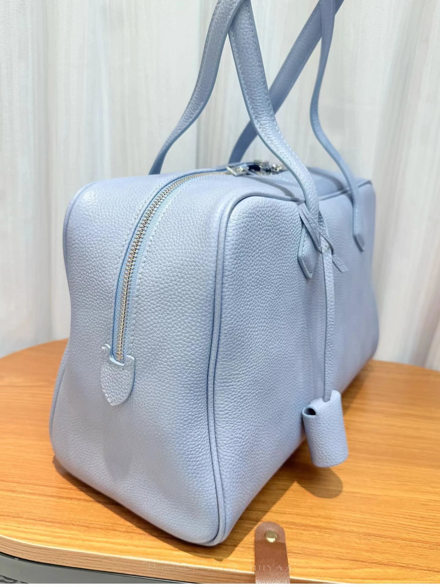 A light blue pebbled leather duffel bag with dual top handles and a side zipper, resting on a wooden surface against a light curtain background.