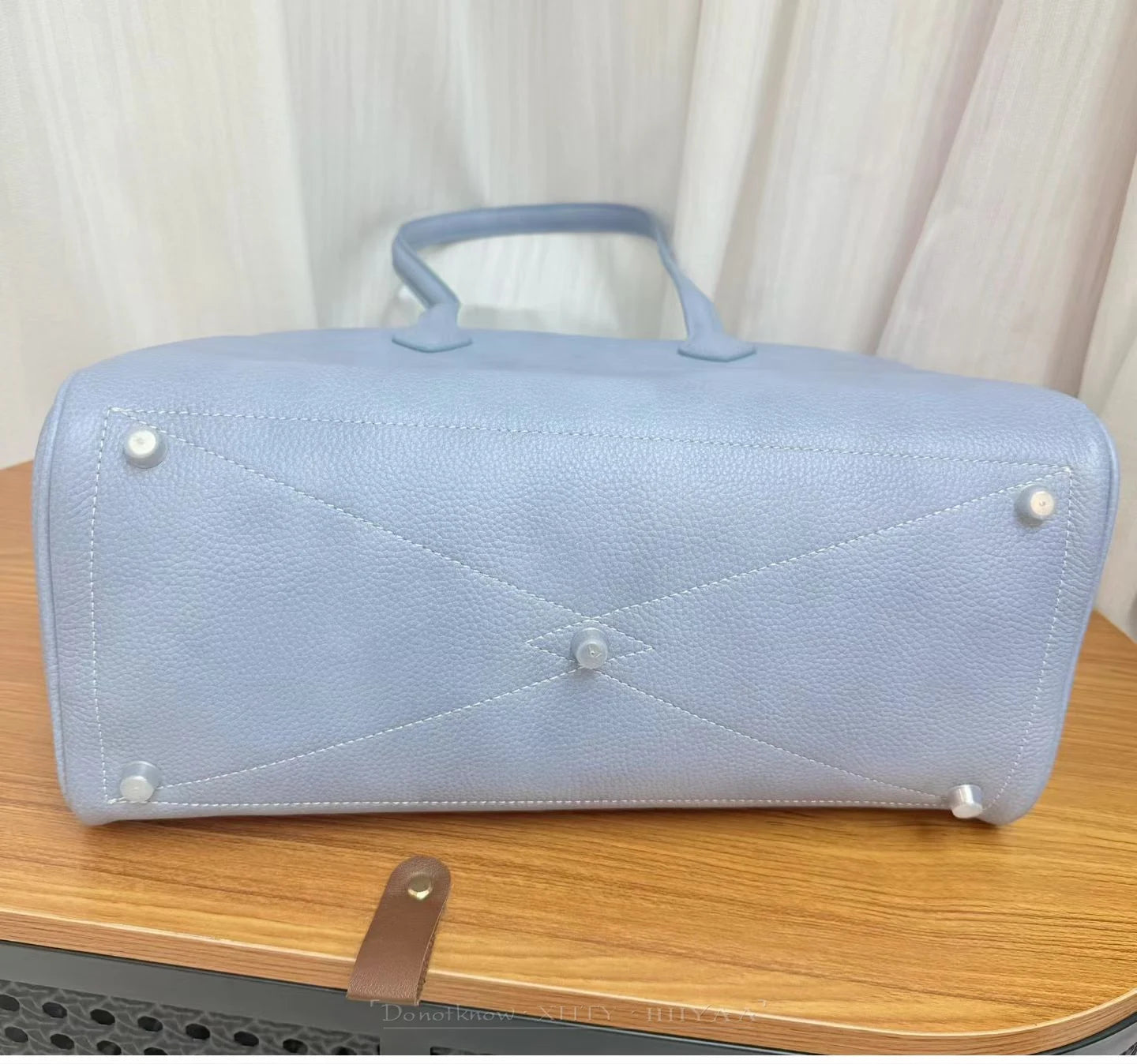 A light blue pebbled leather handbag shown from the bottom, resting on a wooden table with a cream curtain background.