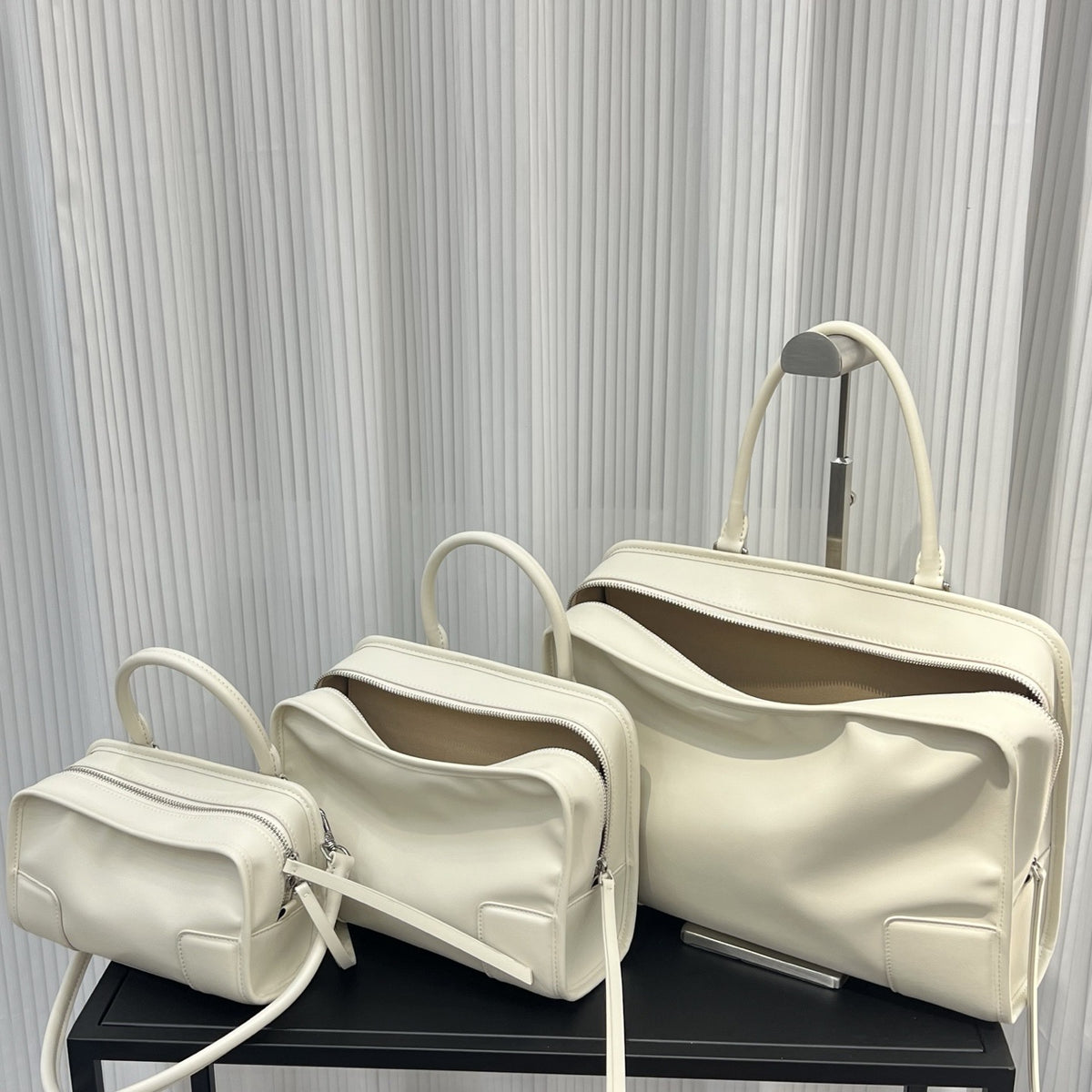 Set of three cream-colored handbags on a black surface with a gray pleated curtain background