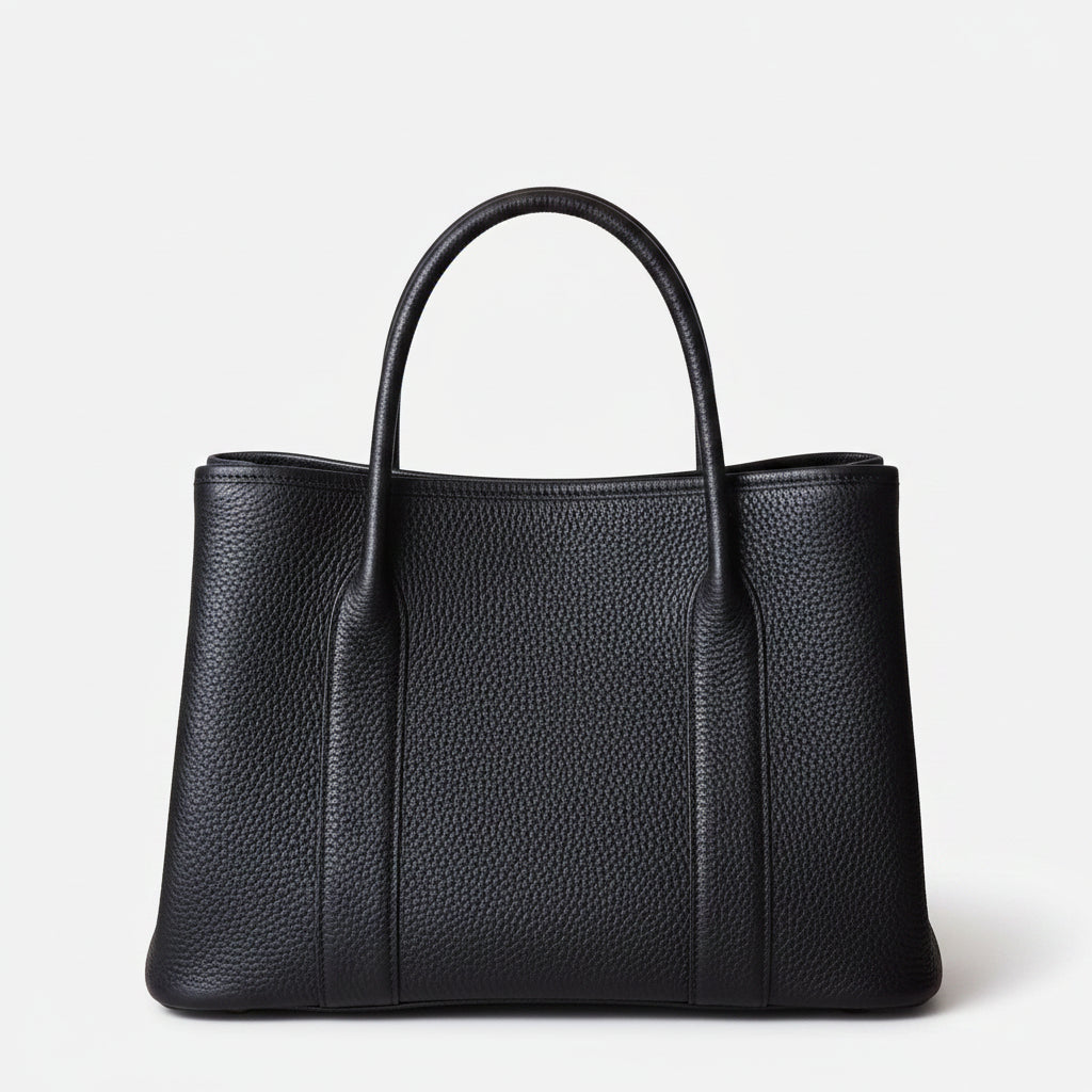 A black leather tote bag with rolled top handles and a small white detail on the top, placed on a wooden surface with a textured wall in the background.
