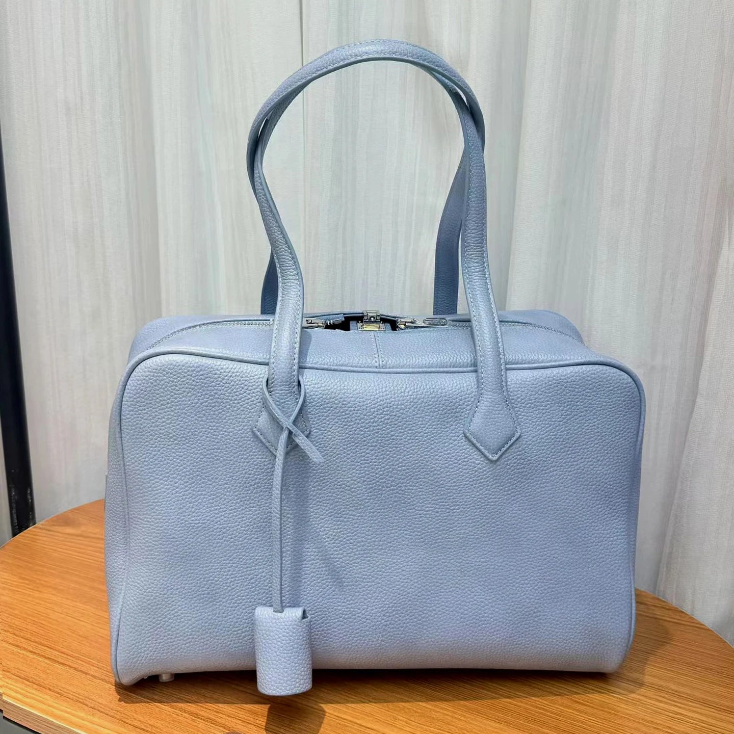 A light blue pebbled leather handbag with dual top handles and a hanging clochette, displayed on a wooden surface against a light curtain background.