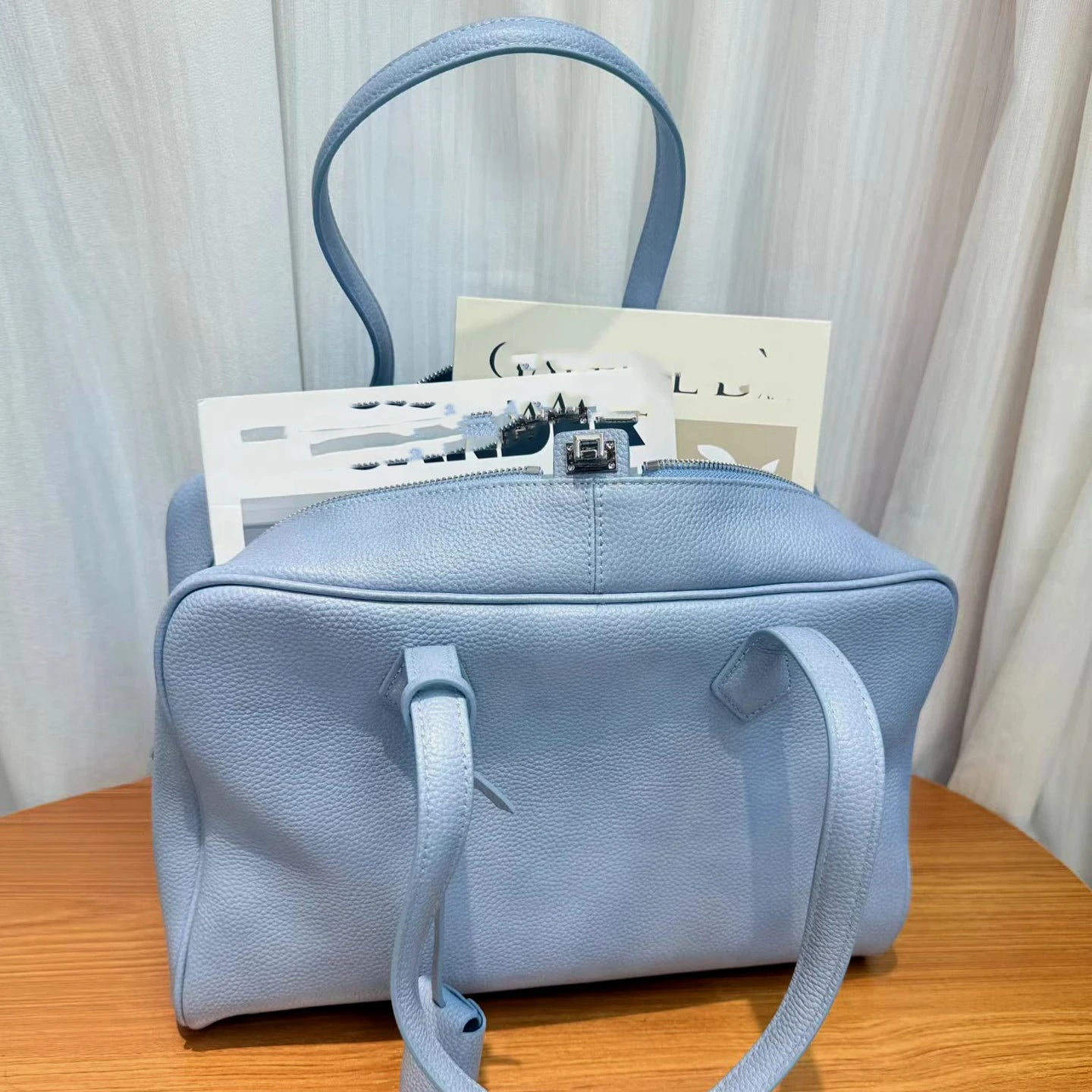 A light blue pebbled leather handbag with dual top handles and a shoulder strap, resting on a wooden surface in front of a white curtain backdrop.