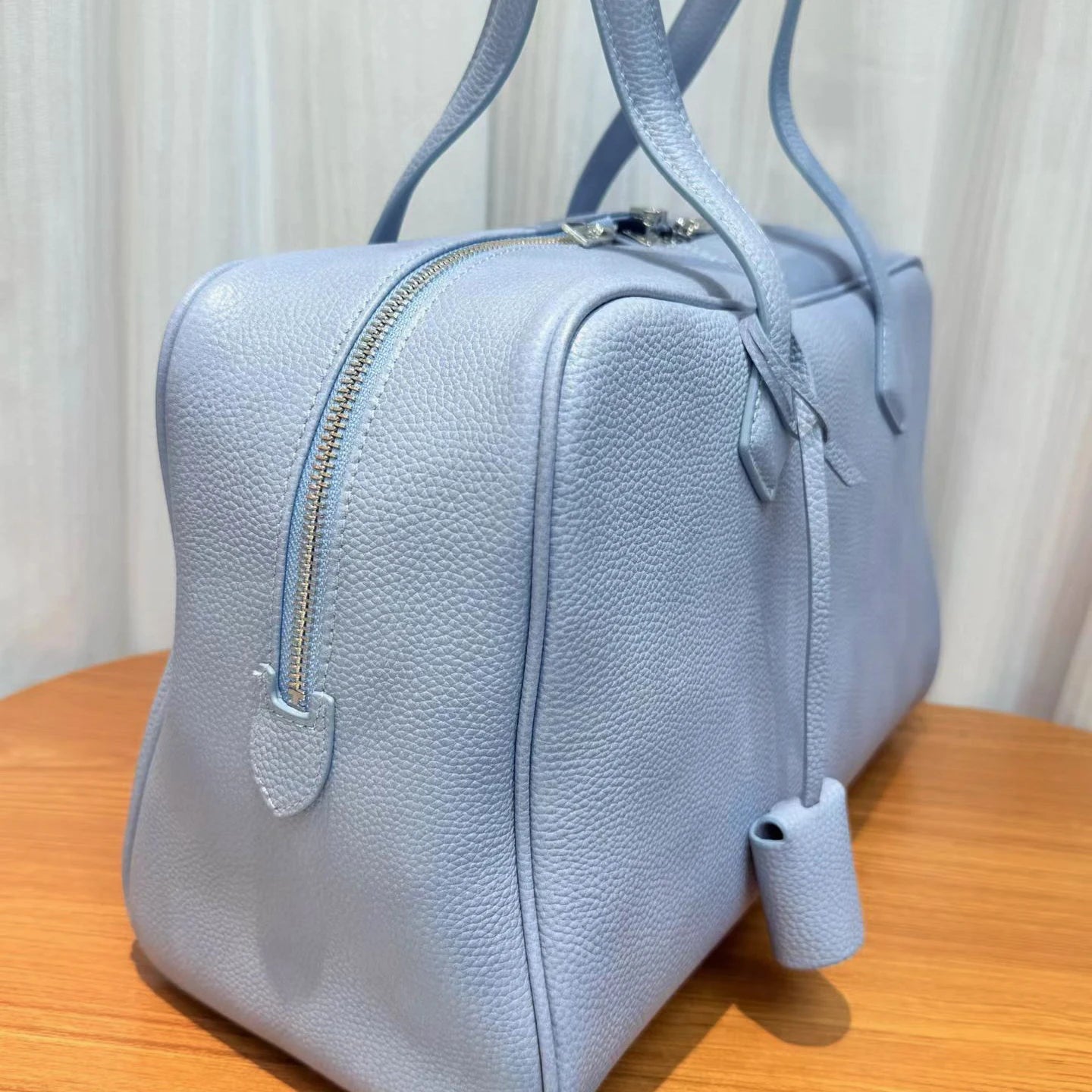 A light blue pebbled leather handbag with silver-tone hardware, shown from a side angle on a wooden surface with a soft-focus curtain background.
