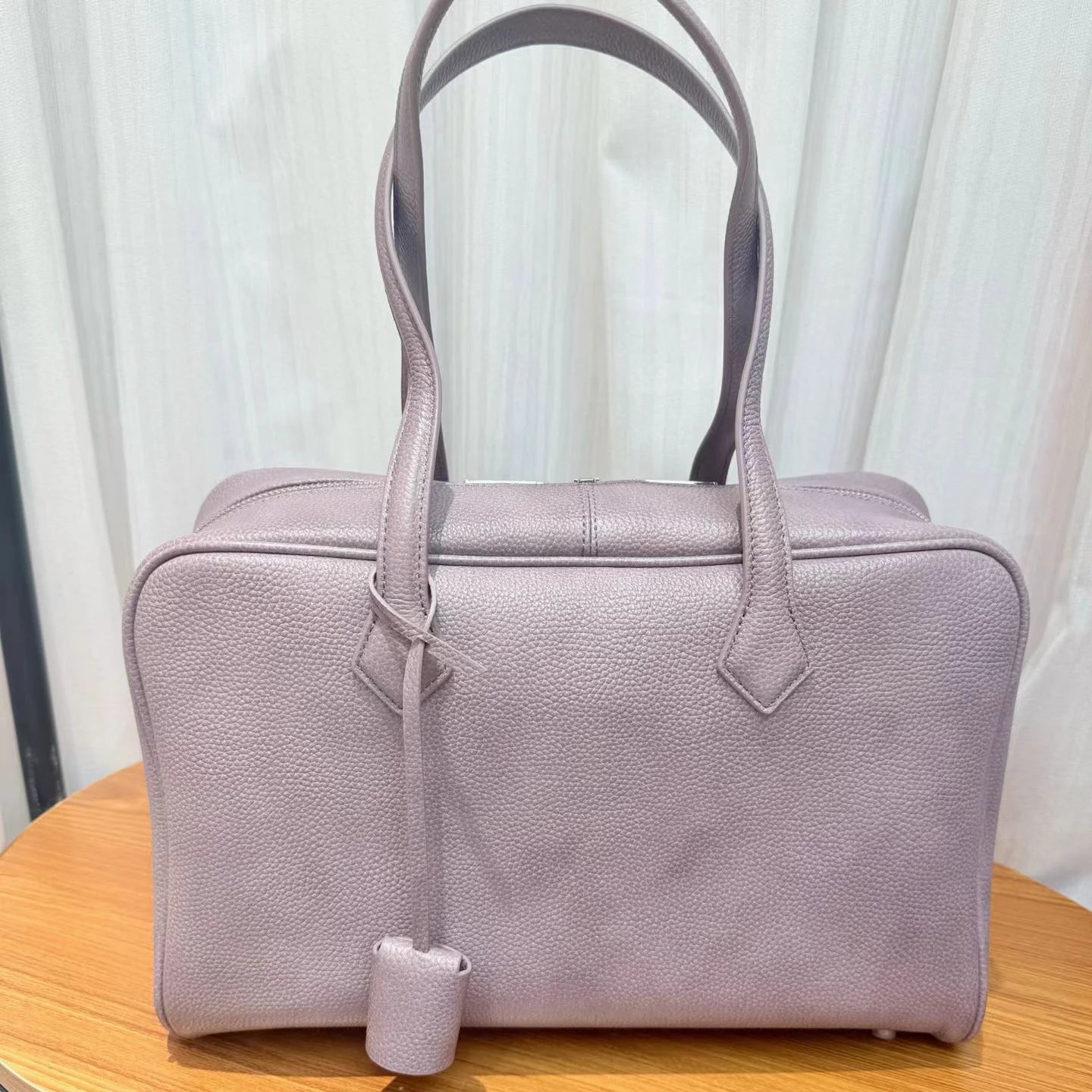 A light purple pebbled leather handbag with double top handles and a small hanging clochette, resting on a wooden surface in front of a white curtain background.