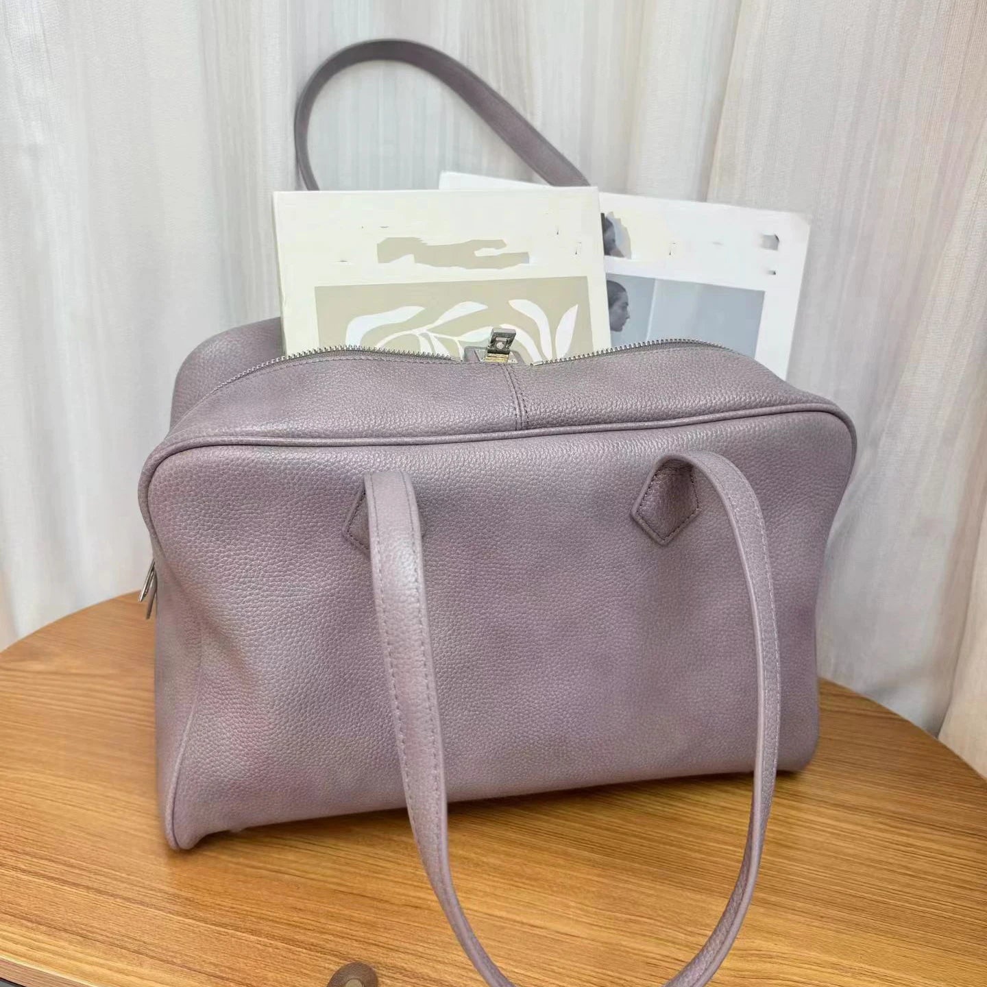A dusty purple pebbled leather travel bag with top handles, shown sitting on a wooden table with travel documents peeking out from the top zipper.