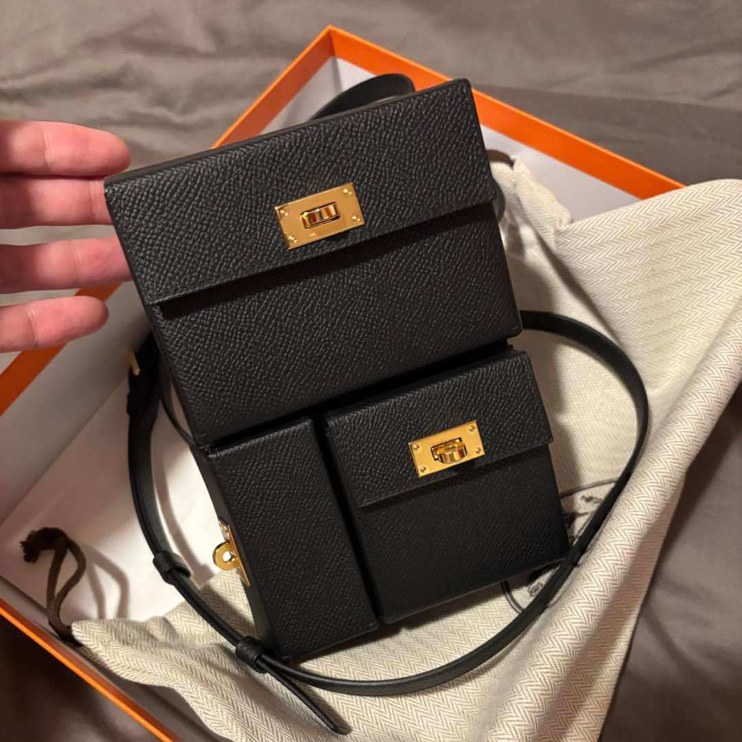 A black textured leather Hermès handbag with gold-tone hardware, resting inside its orange and white Hermès box on a beige dust bag. A hand is visible holding the bag on the left side.