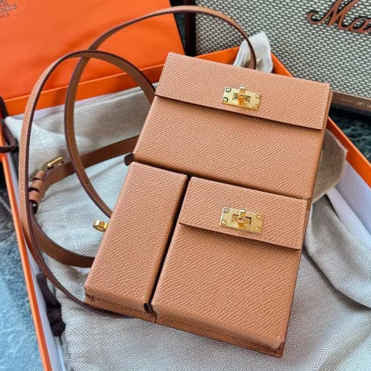 A small structured tan leather handbag with a flap closure and gold-tone hardware, displayed inside an orange box on a dust bag. A large orange Hermès box is visible behind it, and a Marshall speaker sits in the background.