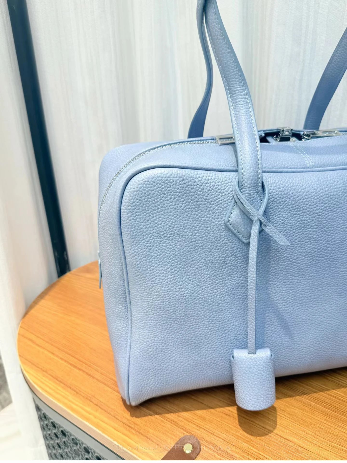 A light blue pebbled leather handbag with dual top handles and a hanging leather tag, resting on a wooden table.
