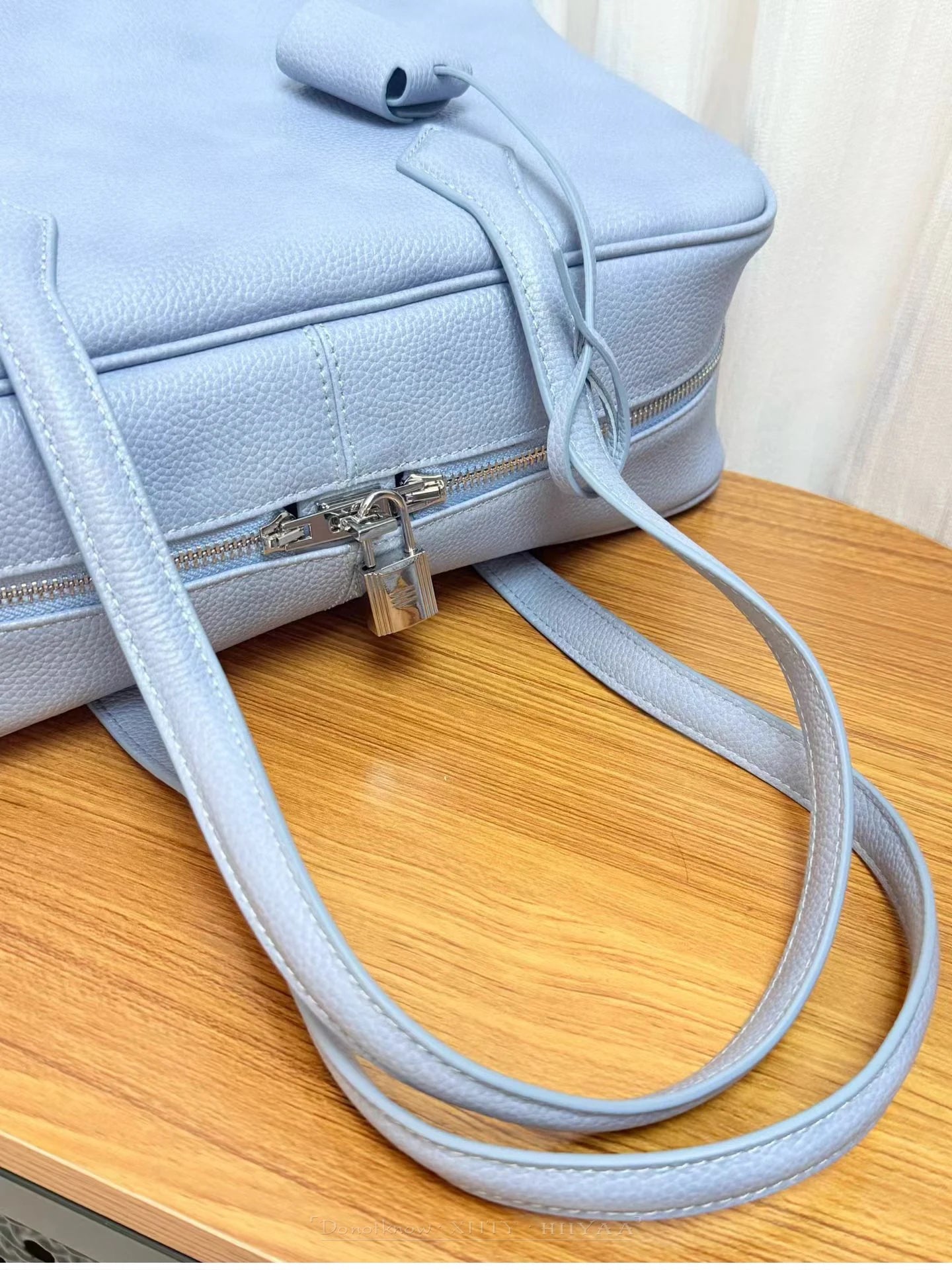 A light blue pebbled leather shoulder bag with a silver padlock clasp and matching strap, resting on a wooden table.