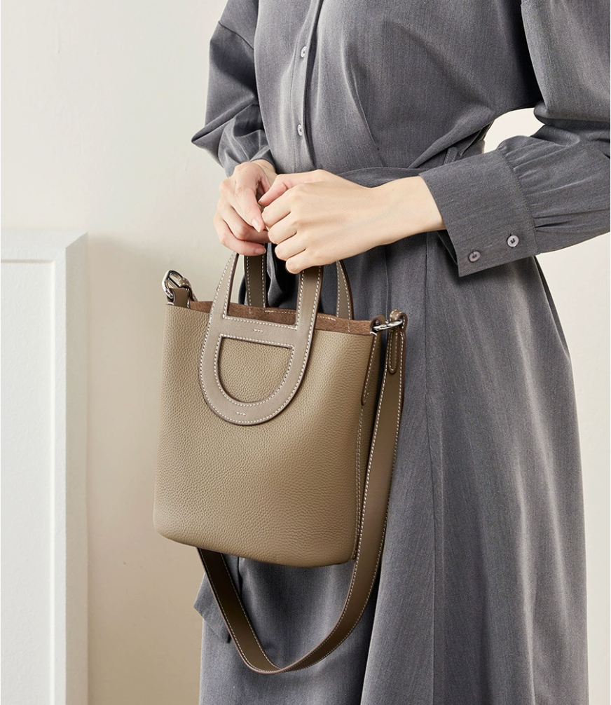 Person holding a beige handbag wearing a gray dress against a neutral background