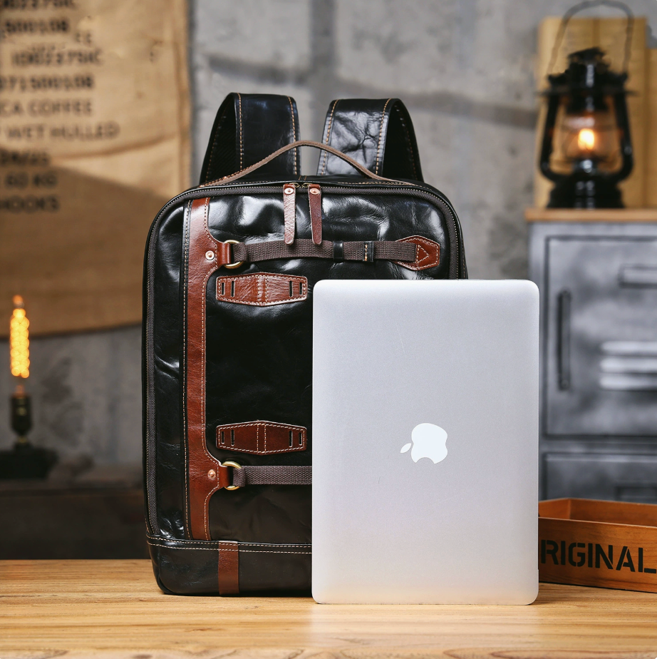Black leather backpack with brown straps next to a silver laptop on a wooden surface.