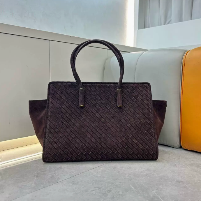 A dark brown woven leather handbag with double top handles and gold-tone hardware, displayed on a light gray floor in front of a modern sofa with white and orange cushions.