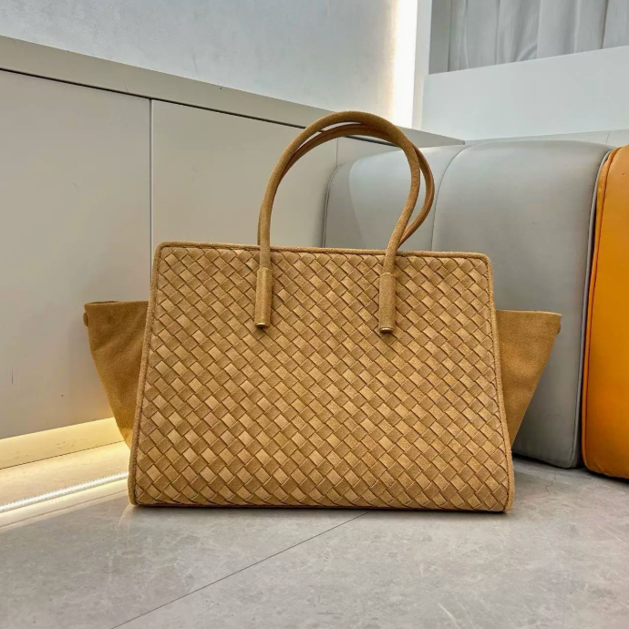 A woven tan leather tote bag with double top handles, displayed on a light gray tiled floor in front of a modern gray and orange sofa.