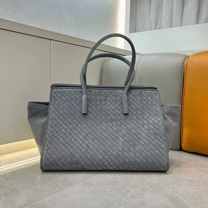 A gray woven leather tote bag with double top handles and gold-tone hardware, placed on a light gray floor in front of a beige sofa with a mustard-yellow cushion.