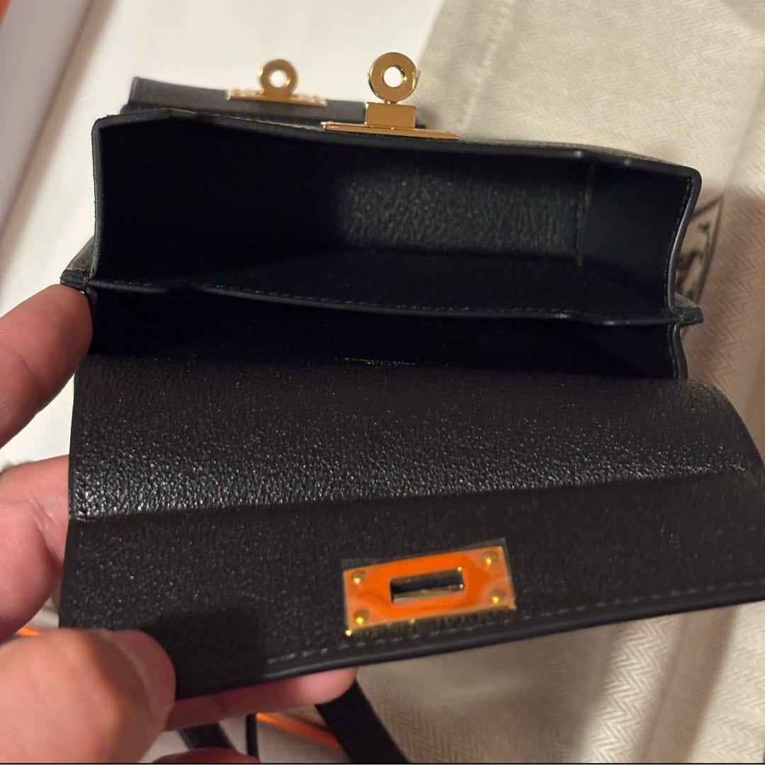 A close-up of a black leather wallet or clutch being held open by a hand, showing a gold-tone clasp and interior. A beige branded dust bag is visible in the background.