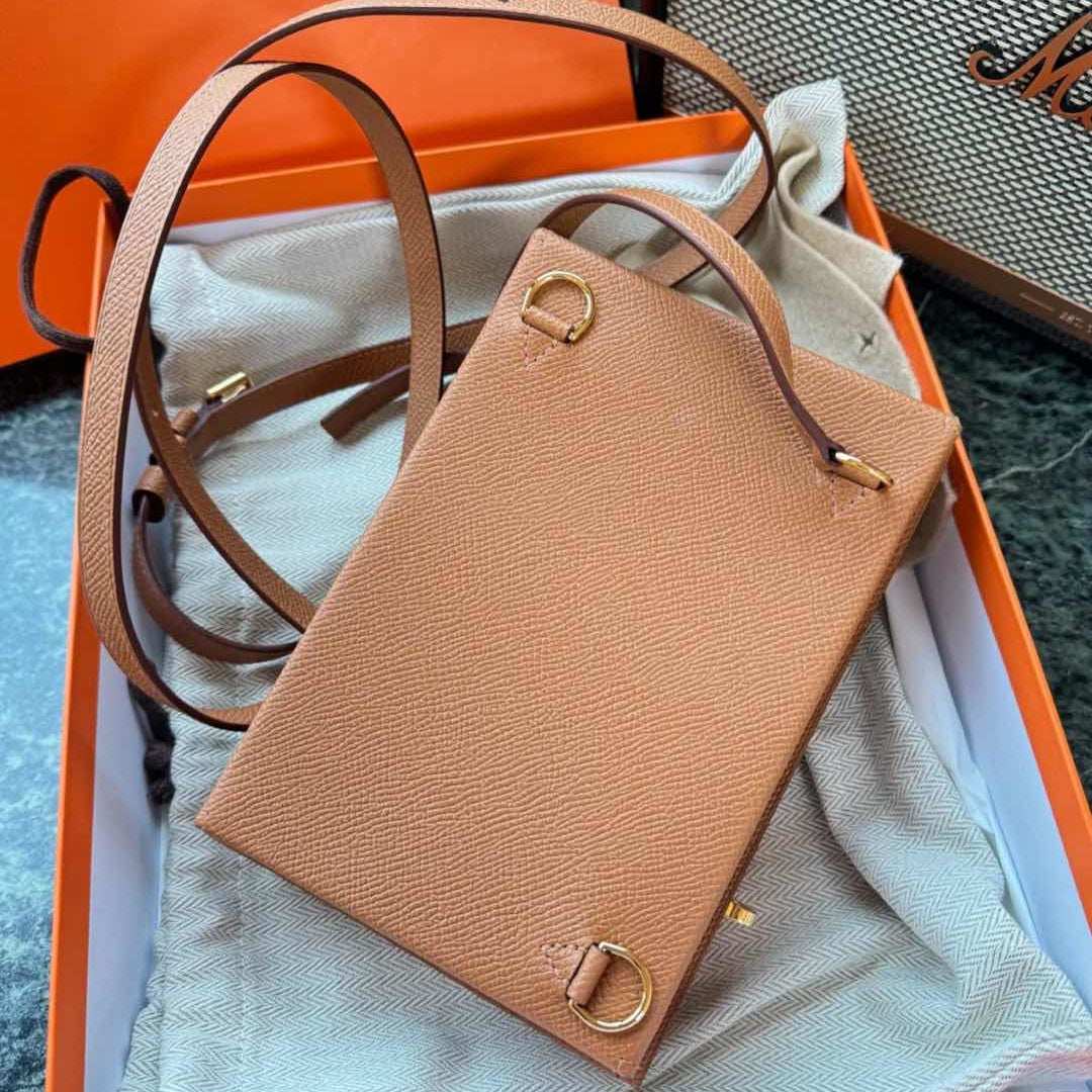 A tan textured leather crossbody bag with gold-tone hardware, presented inside an orange Hermes box with a white dust bag. The Hermes box lid is visible in the background, showing the 'HERMES' logo.